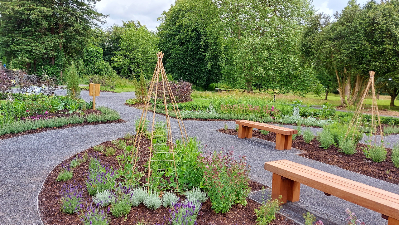 Gardd wedi'i thirlunio gyda llwybrau graean troellog, meinciau pren, planhigion gwyrdd, blodau, a chefnogaeth planhigion pren, wedi'i hamgylchynu gan goed a gwyrddni toreithiog o dan awyr rhannol gymylog. 
